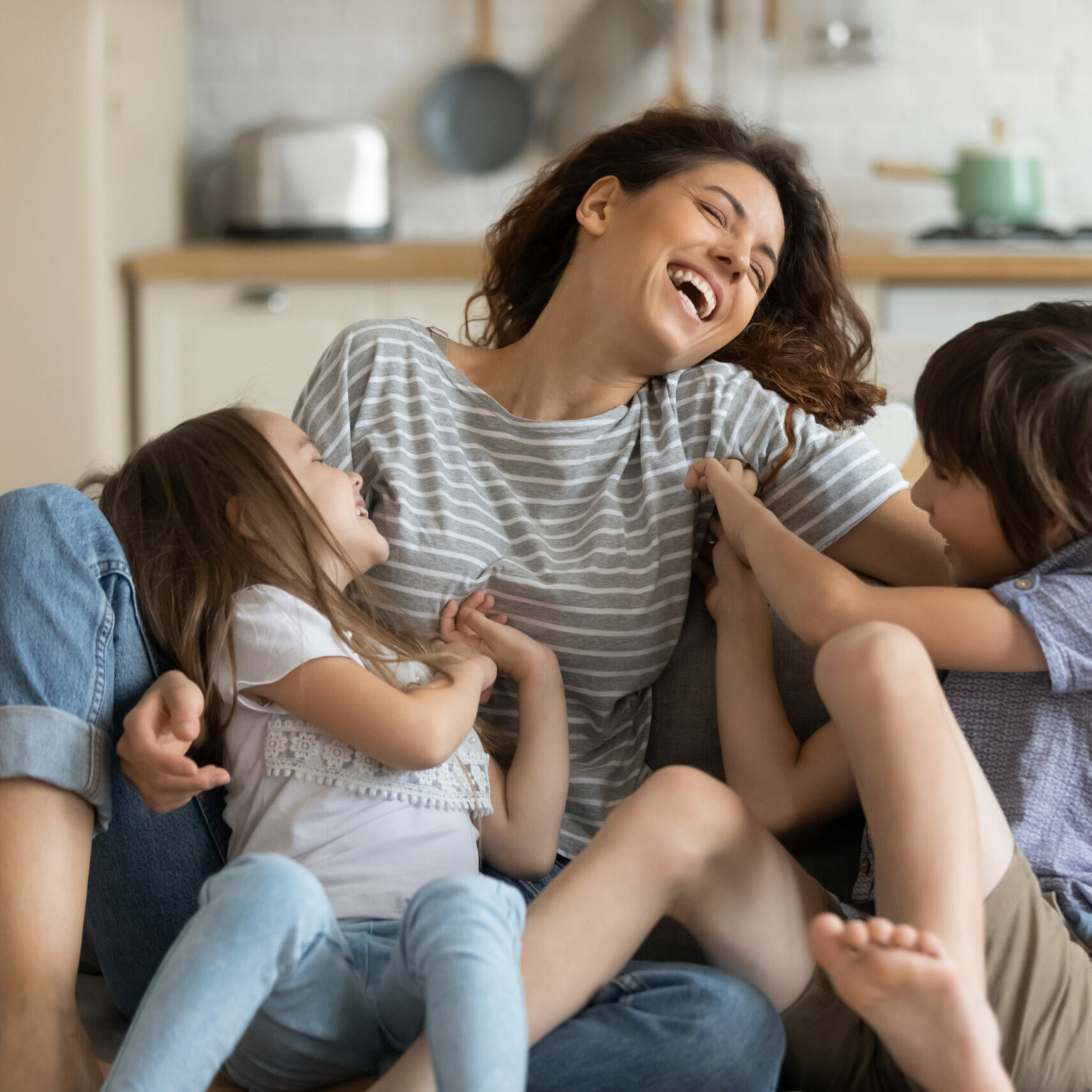 Happy cute daughter and son tickling young attractive mother. Happy cute daughter and son tickling young attractive mother sitting on couch in living room. Close up family having fun at home. Parents playing with cute brother and sister.